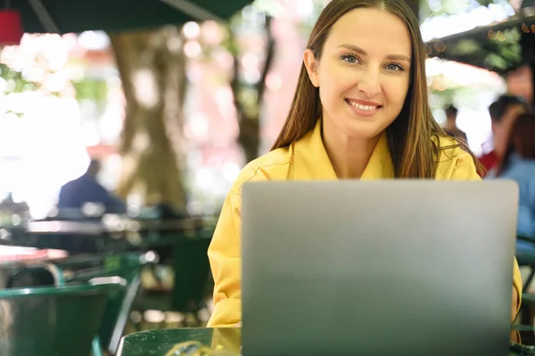 Young freelancer woman using laptop seated outdoors at shaded cafe, suggesting pleasant blend of work and leisure, conveys modern work culture where comfort and productivity meet in informal settings