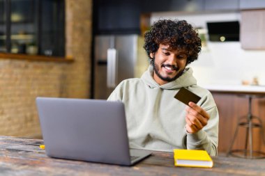 Modern bir mutfakta, rahat bir adam çevrimiçi ödeme sürecine katılır, elinde bir kredi kartı tutarken dizüstü bilgisayarına bakar, dijital işlemlerin kolaylığını ve e-ticareti örnek alır.