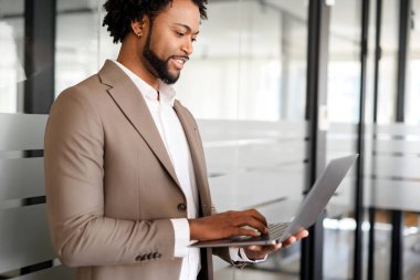 Odaklanmış ve gayretli, çağdaş bir ofis alanının zeminine karşı dizüstü bilgisayarla yazı yazan bir işadamı, üretkenlik ve modern iş yönetimine olan bağlılığını vurguluyor.