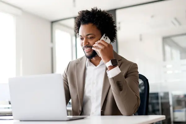 A jovial African-American businessman converses on the phone while working on laptop, highlighting the ability to multitask and maintain client relations in a dynamic office environment
