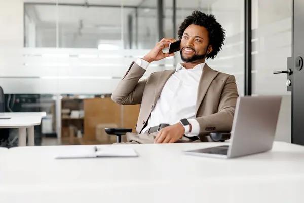 アフリカ系アメリカ人の実業家は 自信とプロフェッショナリズムを除く現代的なオフィス環境で電話会話に従事しています 彼のリラックスした姿勢と笑顔はポジティブな対話を示唆しています — ストック写真