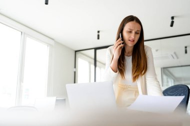 Profesyonel bir iş kadını, bir belgeyi incelerken endişeli bir ifadeyle telefonda konuşur ve iş dünyasındaki karar verme ve sorun çözme konularının zorlu yönlerini yansıtır.