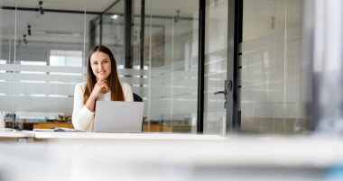 Genç bir iş kadını, modern ofis ortamında masasında oturmuş kameraya dostça bir gülümseme ile bakıyor ve bir dizüstü bilgisayar kullanıyor, cam çevreler çağdaş kurumsal ortamı yansıtıyor.