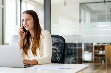 Odaklanmış bir iş kadını, modern ofis arkaplanı ve geniş pencereli önemli iş konuları tartışılırken, dizüstü bilgisayarını kullanırken cep telefonuyla konuşuyor.
