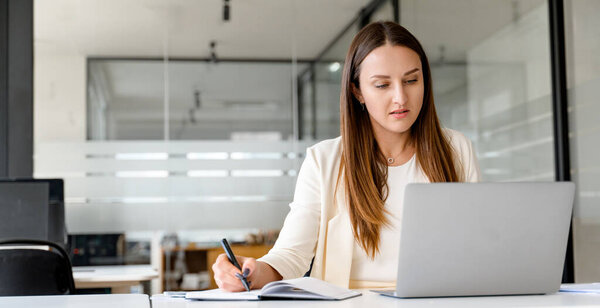 Запечатленная в момент концентрации, молодая деловая женщина внимательно работает над своим ноутбуком, ручкой в руке, символизируя активное участие и вдумчивое принятие решений в современном офисном пространстве