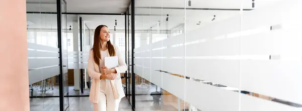 象牙のスーツの陽気なビジネスマンは モダンなガラスのオフィス廊下を歩き ラップトップを運び 彼女の笑顔は 成功した会議や仕事を示唆しています パノラマビュー バナー — ストック写真