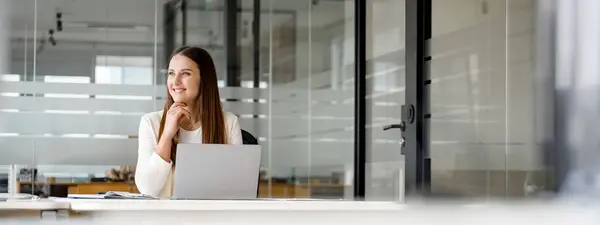 A contemplative businesswoman enjoys a moment of reflection while working on a laptop, her gaze suggesting strategic thought and planning in a spacious office setting. The panoramic view, banner