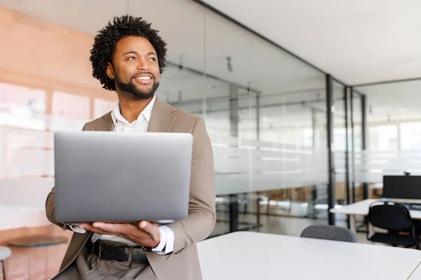 Poised African American businessman in a taupe suit holds his laptop and looks off into the distance, reflecting confidence and vision in a modern office setting standing in bright and open workspace