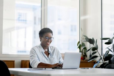 Ofiste dizüstü bilgisayar kullanan Afrikalı siyahi bir iş kadını. Gülümseyen orta yaşlı bayan ofis çalışanı dizüstü bilgisayara bakıyor başarılı kadın girişimci.