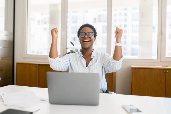 Overjoyed excited african-american woman celebrating victory sitting in the coworking space in front of the laptop, lucky businesswoman screaming yes, receved good news, job offer