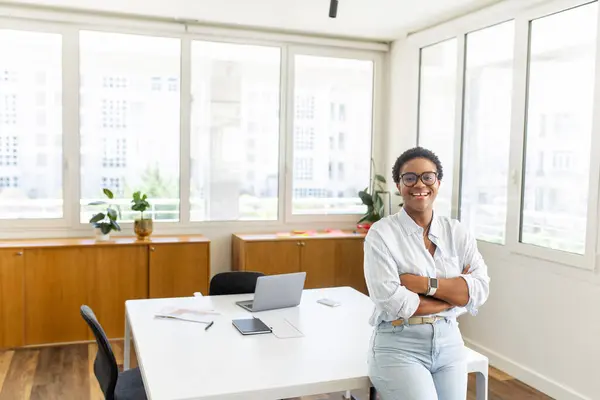 Successful african-american female entrepreneur stands and looks at the camera with arms folded, positive smiling freelancer woman in casual shirt in coworking space, ceo, manager, start-up owner
