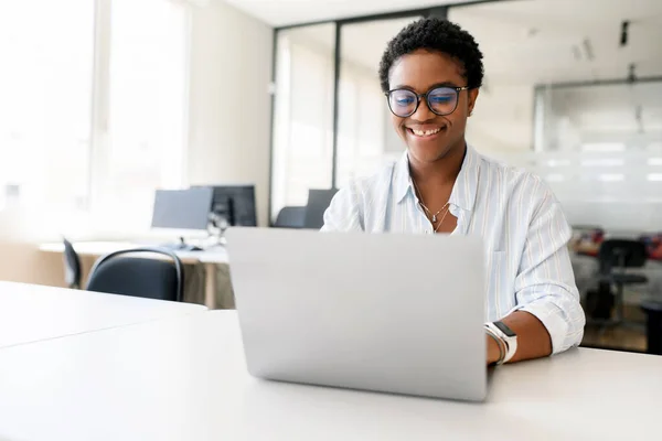 Smiling positive female office employee with computer indoors, African-American woman using laptop for remote work sitting in the modern coworking space, freelancer enjoys work on the distance