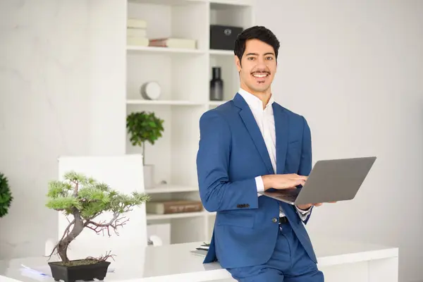 A confident Hispanic businessman in a blue suit stands holding a laptop in a modern office. His poised stance and pleasant smile suggest a successful professional ready for corporate challenges