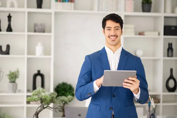 In a modern office setting, a Hispanic businessman expertly navigates his tablet, looking at the camera with a bright smile standing in modern office