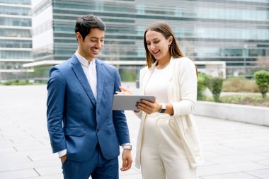 Hispanik bir işadamı ve meslektaşı, ortak bir başarı anında, bir tablet, somutlaştırıcı takım çalışması ve kurumsal bir ortamda işbirliği, arka planda iş binaları gibi içeriklere bakıyorlar.