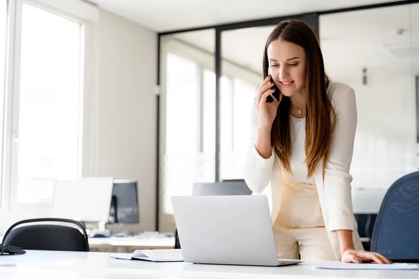 Young businesswoman in a dynamic work pose, speaking on the phone and using her laptop, which highlights her ability to handle multiple tasks efficiently in a fast-paced work environment