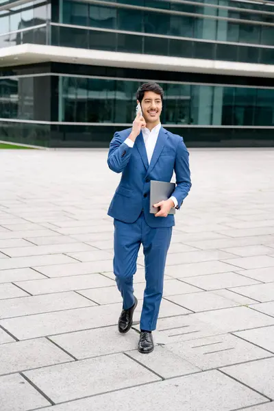 Cheerful Hispanic entrepreneur walks and talks on his phone, his laptop in hand, outside a modern office building, portraying a dynamic and successful professional lifestyle, multitasking and mobility