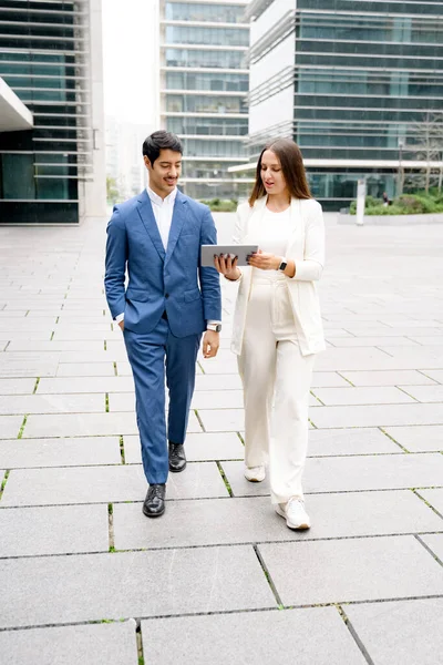 A man and woman, dressed in business attire, engage in a light-hearted conversation while strolling and looking at a tablet, exemplifying a collaborative work moment outside the office.