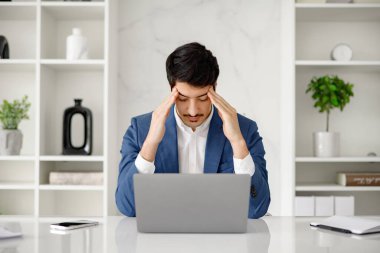 İspanyol bir işadamı dizüstü bilgisayarı üzerinde çalışırken stres ve baş ağrısı yaşıyor ve ofis çalışmalarının zorluklarını anlatıyor. Fotoğraf, işle ilgili stres ve sorun çözme kavramını temsil ediyor.