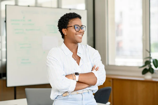 アフリカ系アメリカ人の実業家が ノートパソコンで近代的なオフィスに立って 目をそらす 腕が交差した短い髪型の野心的な笑顔の多民族マネージャーの従業員の女性 — ストック写真