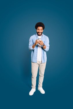 A cheerful young man with an afro hairstyle is holding a smartphone with both hands and smiling at the screen. The concept highlights modern connectivity and the joy of social interaction. clipart