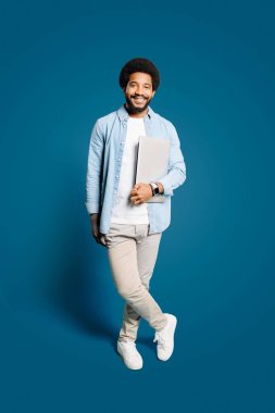 Günlük kıyafetlerle gezen neşeli genç bir adam dizüstü bilgisayar tutuyor, mavi bir arkaplana karşı duruyor. Görüntü canlı, profesyonel ama yaklaşılabilir bir titreşim yakalar, teknoloji veya eğitim içeriği için idealdir.