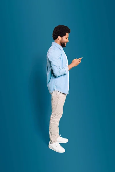 彼の携帯電話に深く関わった若い男性は 現代生活におけるモバイルデバイスのユビキタスな性質を示しています ブルーで隔離されたスマートフォンを使用したブラジルの男性の全長写真 — ストック写真