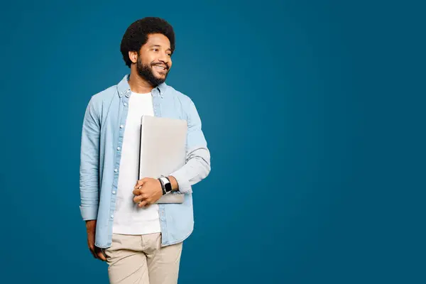 A smiling Brazilian young man, freelancer or student carrying a laptop standing isolated on blue, exuding confidence and readiness. Concept of readiness for business or learning opportunities.