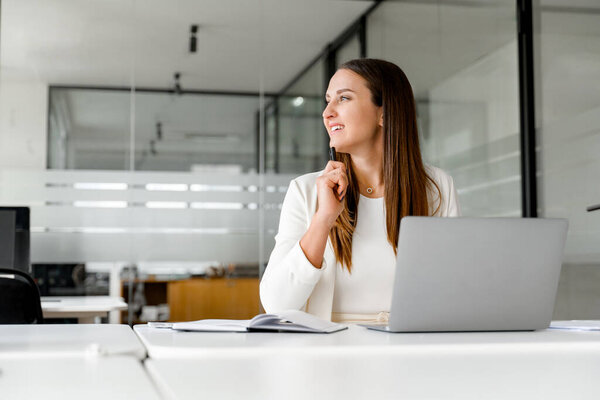 A businesswoman ponders with a smile, looking away from the laptop, contemplating a new idea, or reflecting on a successful deal. Concept of creative and strategic aspects of business thinking