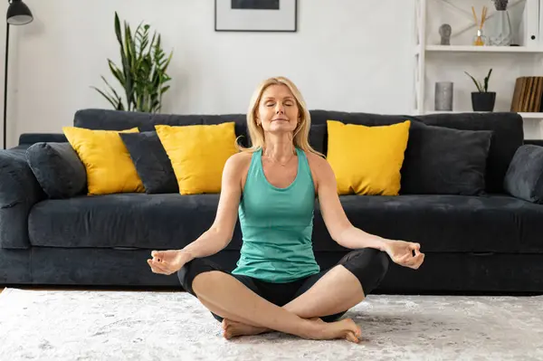 Training at home. Mature woman in a easy pose sits on the floor in sportswear barefoot and with closed eyes. Relaxing antistress yoga at home concept