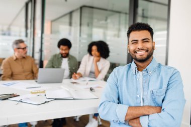 Kot gömlekli neşeli Hintli profesyonel kollarını kavuşturmuş, parlak bir gülümsemeyle arka planda birlikte çalışarak, pozitif ve enerjik bir ofis kültürünü temsil ediyor.