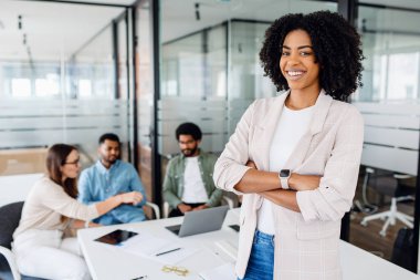 Modern bir ofis ortamında kollarını kavuşturup arka planda iş birliği yapan takım sinerjisi ile neşeli bir gülümsemesi olan zeki, yüksek yetenekli Brezilyalı genç kadın.