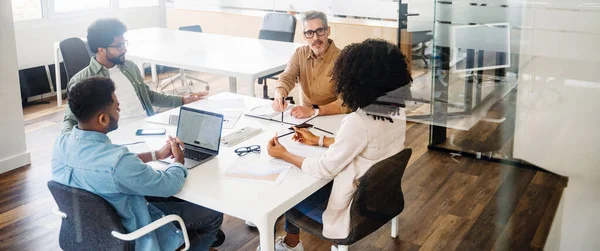 In a wide office shot, four professionals are deep in discussion around a table, showcasing a dynamic and collaborative team in action. A diverse team in a friendly and inclusive workplace atmosphere