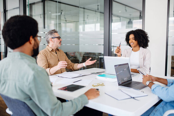 Colleagues engage in a lively exchange of ideas in an office, with a mature man, leader or mentor, in glasses gesturing to emphasize a point, embodying a collaborative and engaging work discussion.