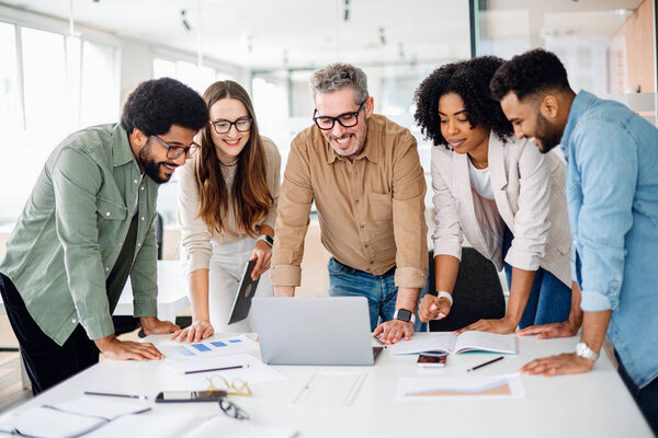 Различные коллеги собираются вокруг ноутбука с яркими выражениями, что свидетельствует об успешном совместном усилии и живой мозговой штурм сессии в хорошо освещенном, современном рабочем пространстве, концепции командной работы