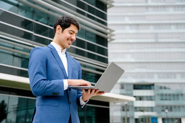A smiling Hispanic businessman stands with an open laptop in front of a modern office, embodying the concept of accessible technology and business efficiency in todays fast-paced world.