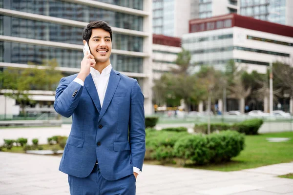 This image captures a Hispanic businessman mid-conversation on his phone, the focused expression and modern city backdrop reflecting his active role in the fast-paced business world.