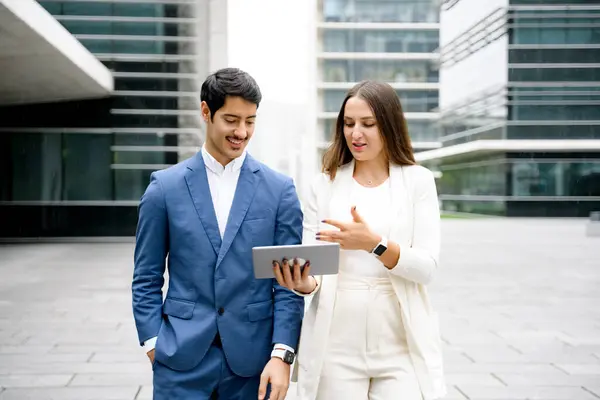 In a casual business exchange, a man and woman walk while discussing a project on a digital tablet, epitomizing on-the-go professionalism in an urban setting.