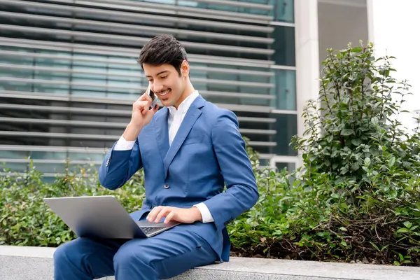 A business-savvy Hispanic male utilizes his laptop and smartphone simultaneously, seated in a relaxed outdoor setting, epitomizing the modern mobile office of a contemporary entrepreneur