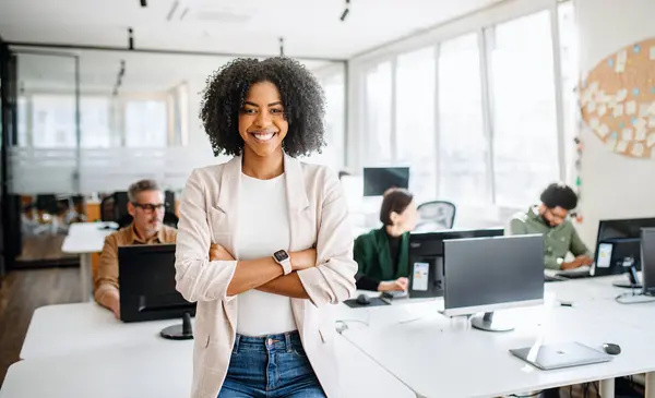 A confident Brazilian female professional stands with arms crossed in an open office environment, her friendly demeanor and business casual wear reflecting a modern and approachable office culture
