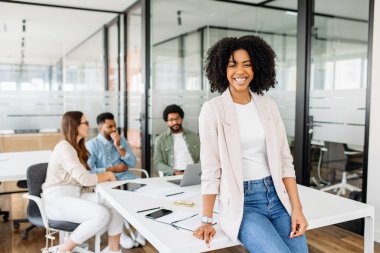 Çekici, zeki Brezilyalı kadın, kadın bir ofis çalışanı, rahat bir şekilde duruyor, ekibi arka planda, rahat ama üretken bir toplantı atmosferini somutlaştırıyor.