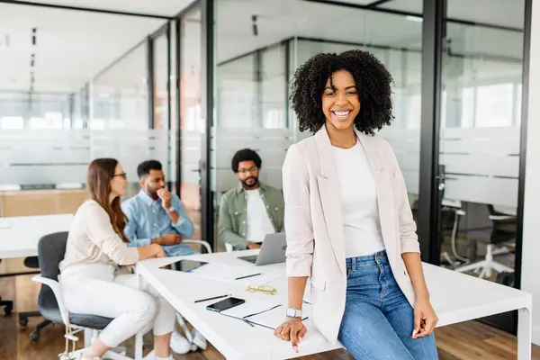 Charming smart Brazilian woman, a female office employee, stands with easy confidence, her team is in the background, embodying a relaxed yet productive meeting atmosphere