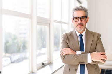 Kıdemli işadamı, iyi ışıklandırılmış, kollarını kavuşturmuş, profesyonellik ve tecrübe dolu bir ofiste güven içinde duruyor. Gri saçları, özel dikim bej takım elbisesi ve kravatı sofistike bir şirket tarzını yansıtıyor.