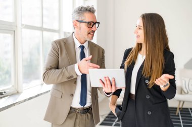 Gri saçlı iş adamı, genç meslektaşı ile yakınlaşıyor ve parlak, modern ofis alanında bir tablet ekranı paylaşıyor. Canlandırılmış konuşmaları ve işbirlikçi duruşları akıl hocalığı kavramının altını çiziyor