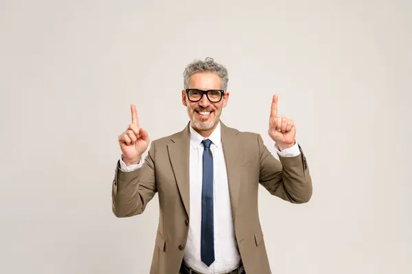 Senior executive points upwards with both hands, grey hair and smart beige suit complimenting his knowledgeable expression. This image evokes concepts of growth, future goals, and visionary leadership