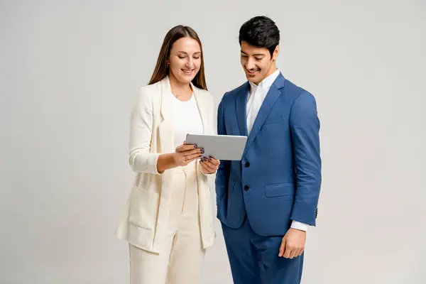 A professional woman and man team, in a beige suit and blue suit respectively, are engaged with a tablet, symbolizing teamwork and collaboration in a modern corporate setting.