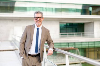 Tecrübeli işadamı korkuluklara yaslanıyor. Duruşu gevşemiş ama kendine güveni tam. Arka planı modern bir bina oluşturuyor. Gelişigüzel profesyonellik ve ulaşılabilirliğin karışımı.