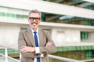 Üst düzey bir yönetici kollarını kavuşturup modern bir ofis ortamının önünde güç ve kararlılık hissi yayıyor. Profesyonel duruşu onu tecrübeli bir lider yapıyor.