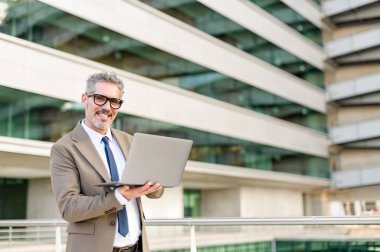Kahverengi takım elbiseli 60 'lı yaşlarda kendinden emin bir işadamı dışarıda dizüstü bilgisayarını büyüleyici bir gülümsemeyle tutuyor, deneyimin ve modern teknolojinin birleşimini gösteriyor.