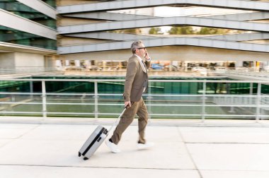 Hareket halindeyken yakalanan bu kıdemli işadamı, bavuluyla hızlı bir şekilde yürürken bir telefon görüşmesi ile meşgul. Çağdaş işlerin dinamik doğasının bir görüntüsü, yan görüş.